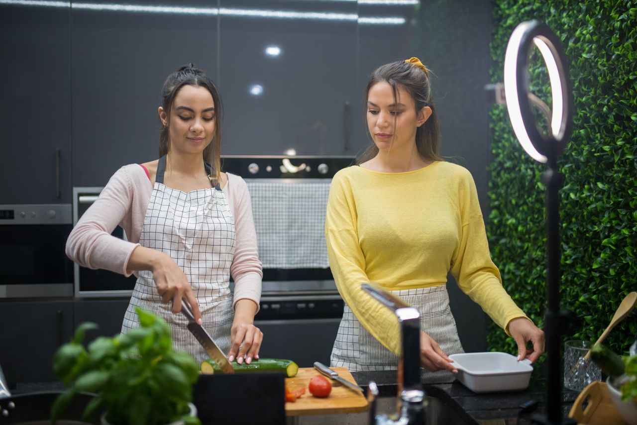 Deux femmes cuisinent et préparent des plats dans une cuisine moderne, éclairées par un ring light. Activité culinaire en collaboration.